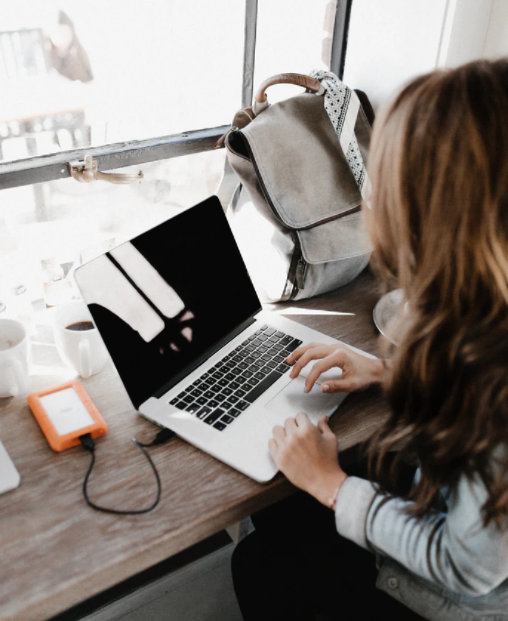 Lady sat at a table in a coffee shop with her bag, Apple Laptop and Hard drive, backing up her data.